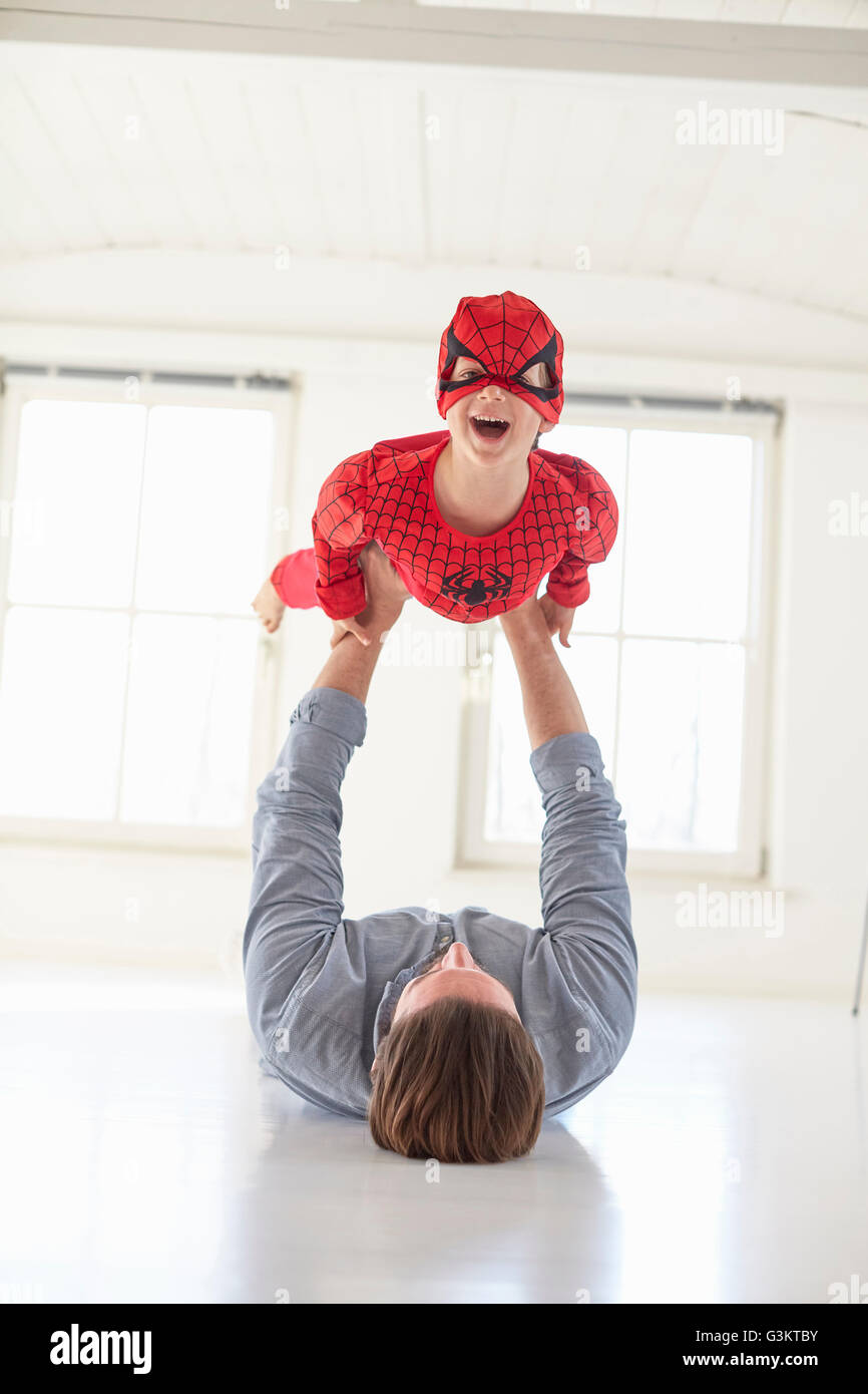 Padre giacente sul piano tenendo premuto fino figlio indossando il costume del supereroe Foto Stock
