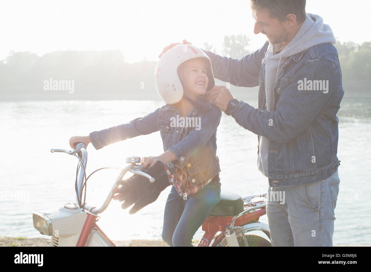 Giovane ragazza seduta sul padre, ciclomotore padre mettendo casco sulla figlia di testa, sorridente Foto Stock