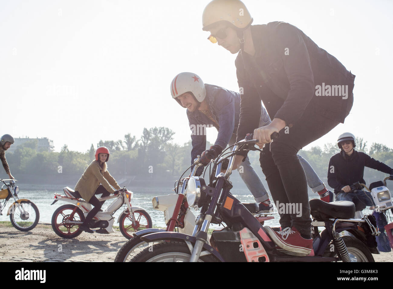 Gruppo di amici in sella ciclomotori accanto al lago, due uomini fare acrobazie Foto Stock
