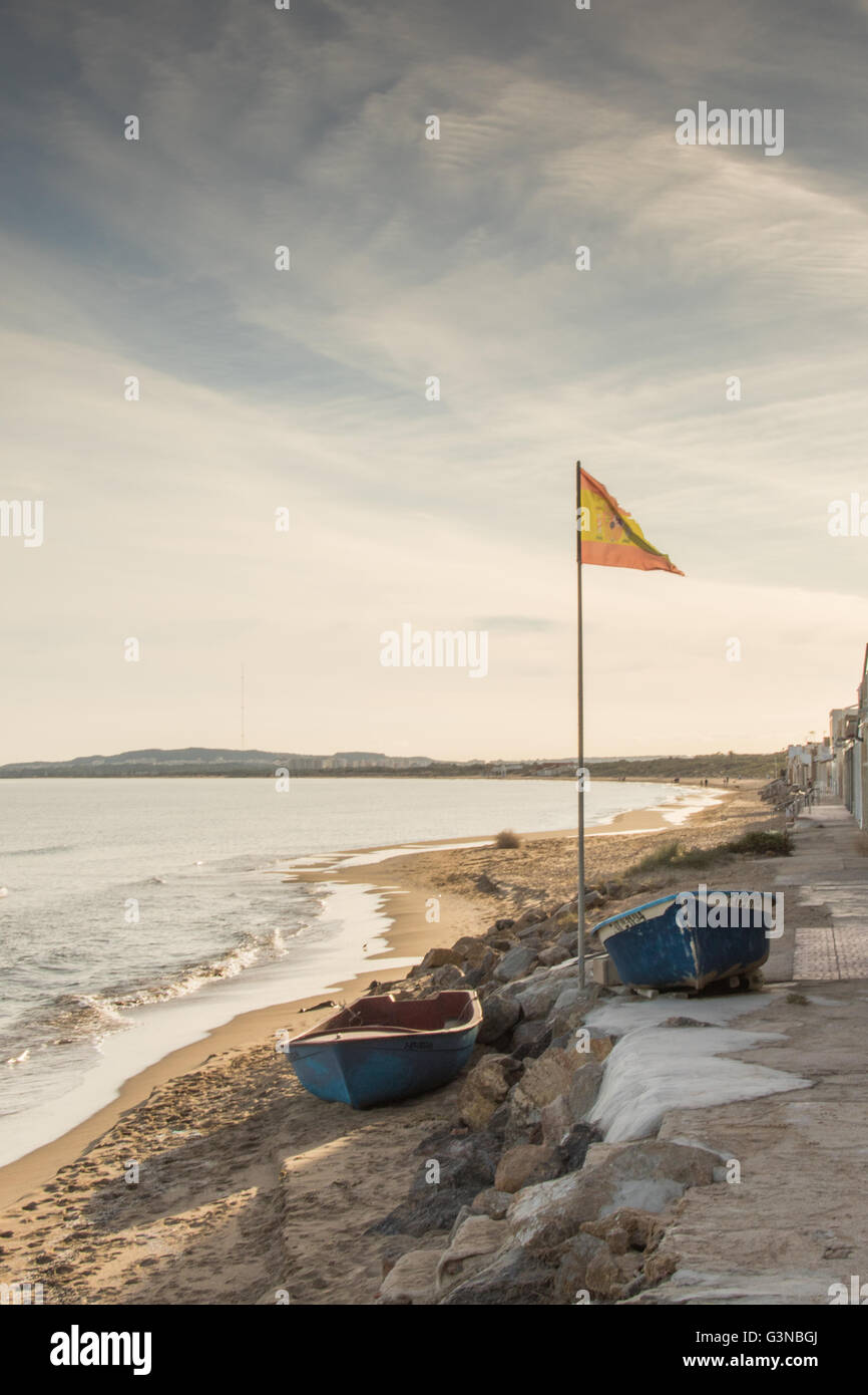 Due piccole imbarcazioni su una piccola spiaggia di sabbia con una bandiera della Spagna soffiando leggermente. Foto Stock