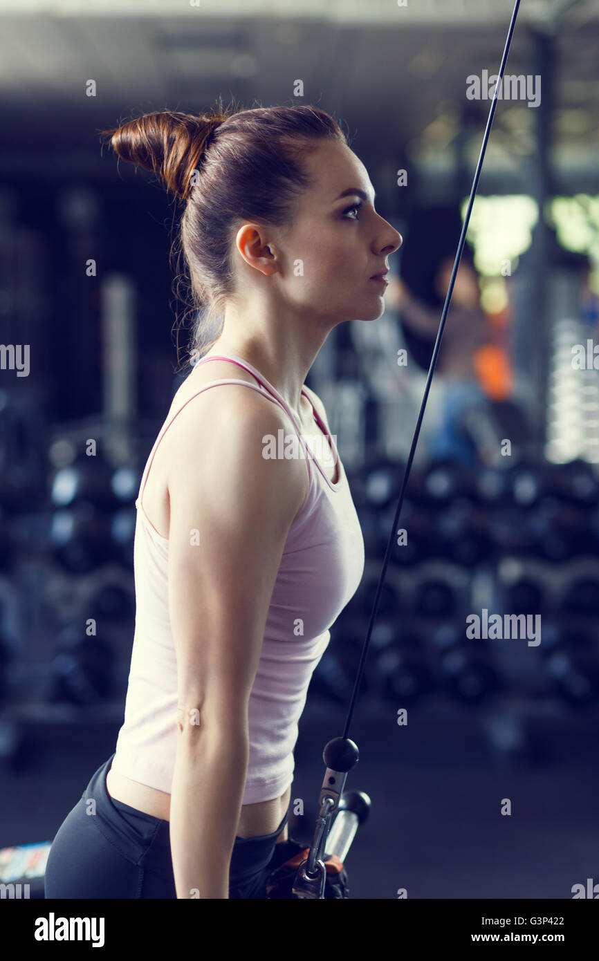 Giovane donna slim facendo pushdown sul cavo macchina in palestra. Atletica formazione ragazza tricipiti nel centro fitness. Immagine dai toni Foto Stock