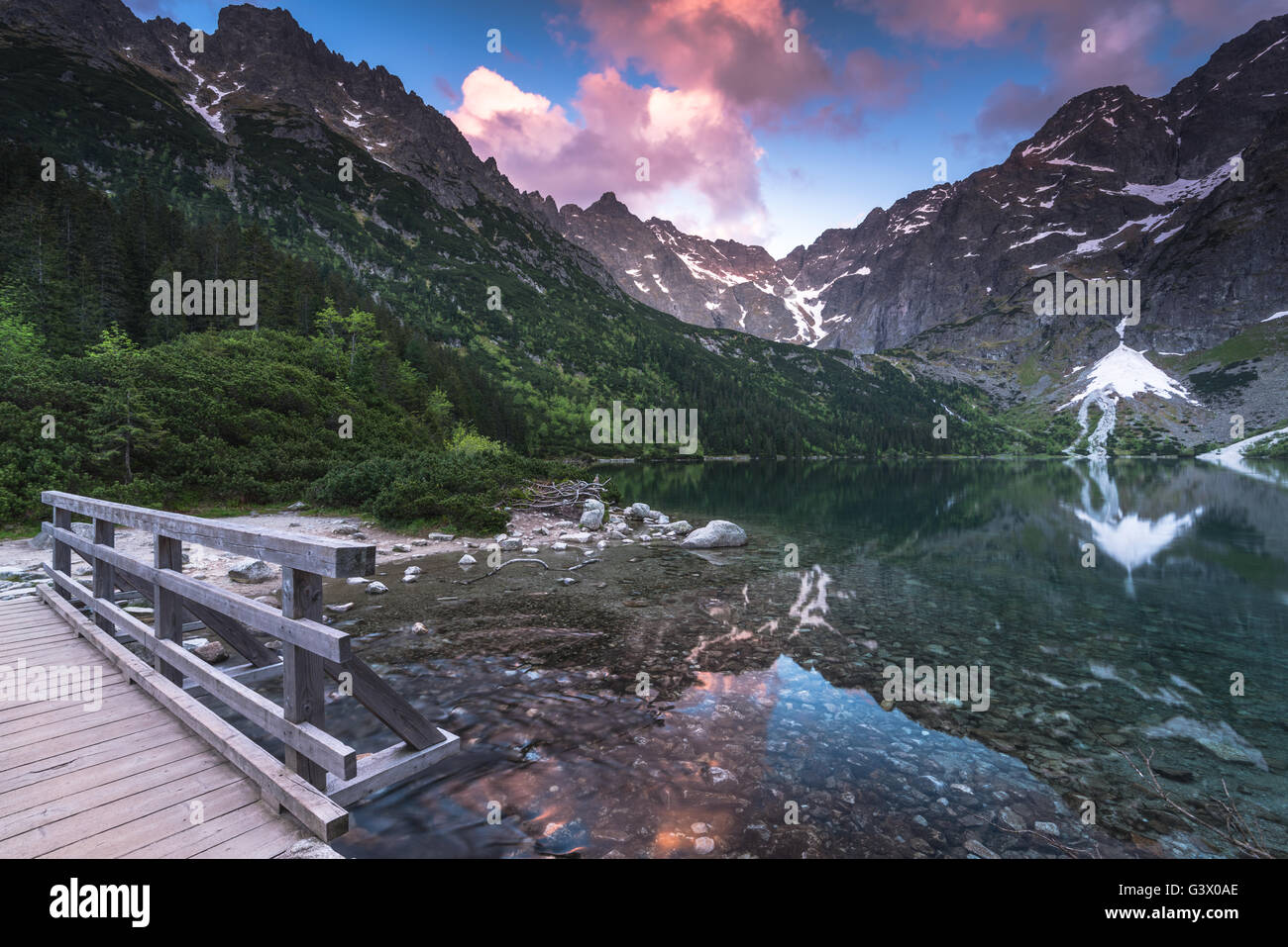 Piedi in legno ponte in hing montagne sopra il lago con drammatica il cielo al tramonto Foto Stock