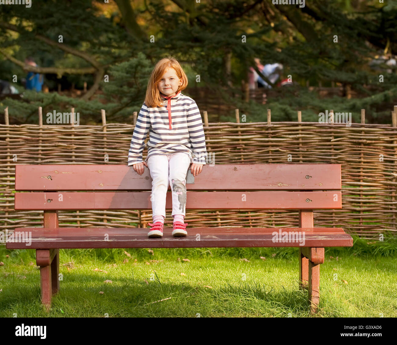 Seduta su una panchina. Una giovane ragazza con i capelli rossi si trova sul retro di una panchina nel parco di fronte alla telecamera. Foto Stock