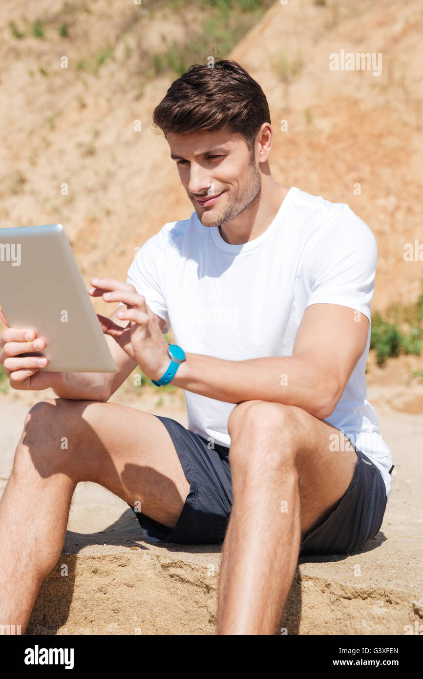 Sorridente giovane uomo seduto e utilizzo di tablet sulla spiaggia Foto Stock Sorridente giovane uomo seduto e utilizzo di tablet sulla spiaggia Foto Stock