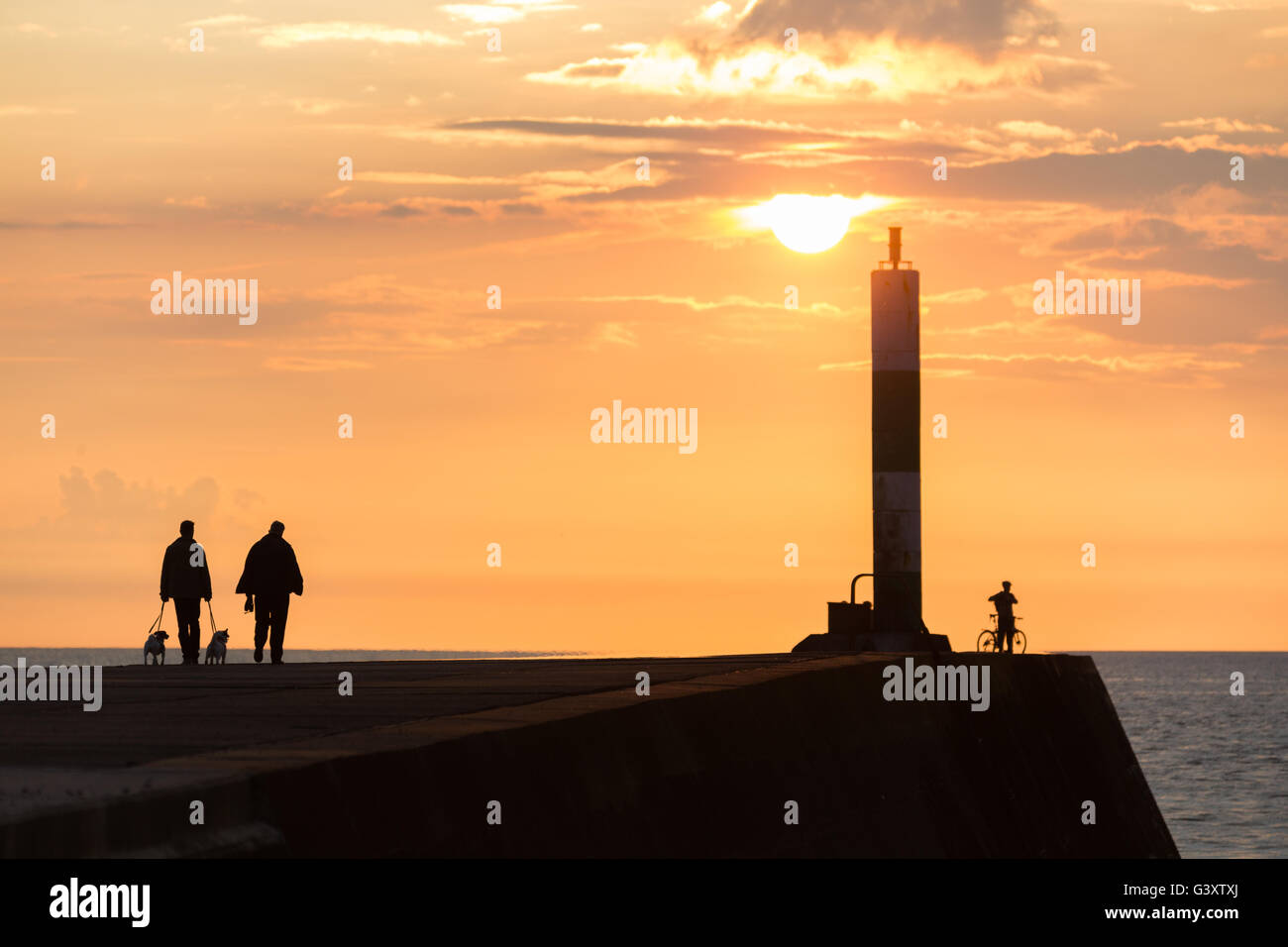 La gente che camminava lungo il mare di calcestruzzo break sull'ingresso a Aberystwyth Harbour come il sole comincia a impostare sulla costa occidentale del Galles. Foto Stock