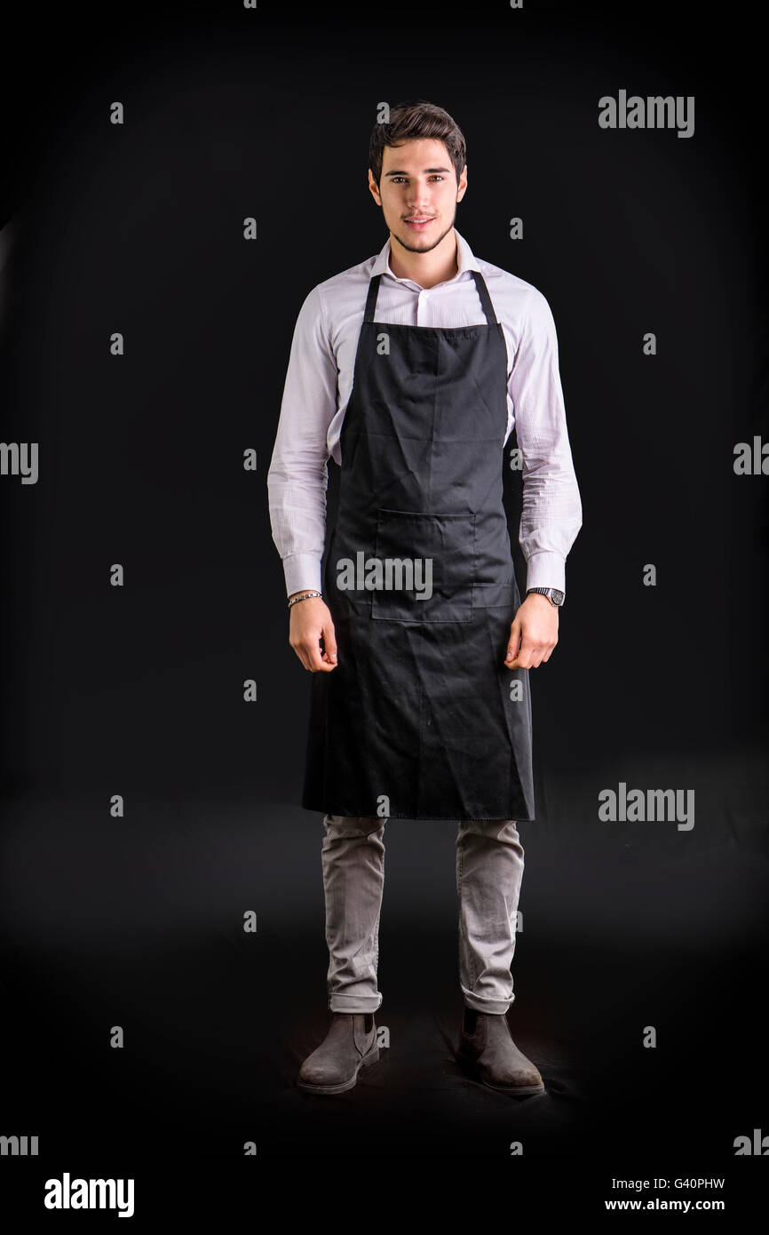 Il giovane chef o il cameriere in posa, indossare un grembiule nero e camicia, su sfondo scuro Foto Stock