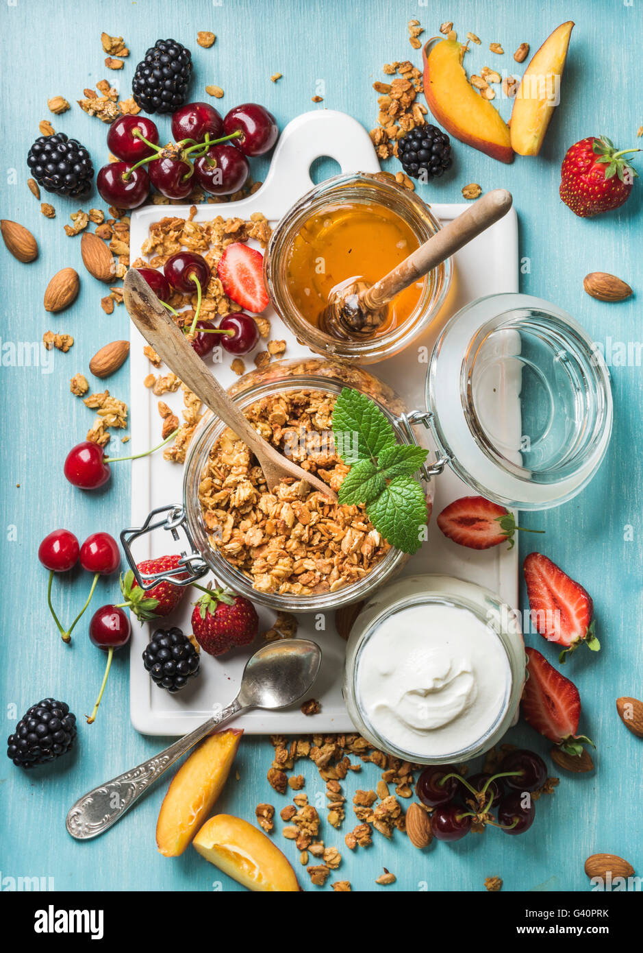Sana colazione ingredienti. Oat granola in aprire il vasetto di vetro, yogurt, frutta, bacche, miele e menta su ceramica bianca su scheda Foto Stock