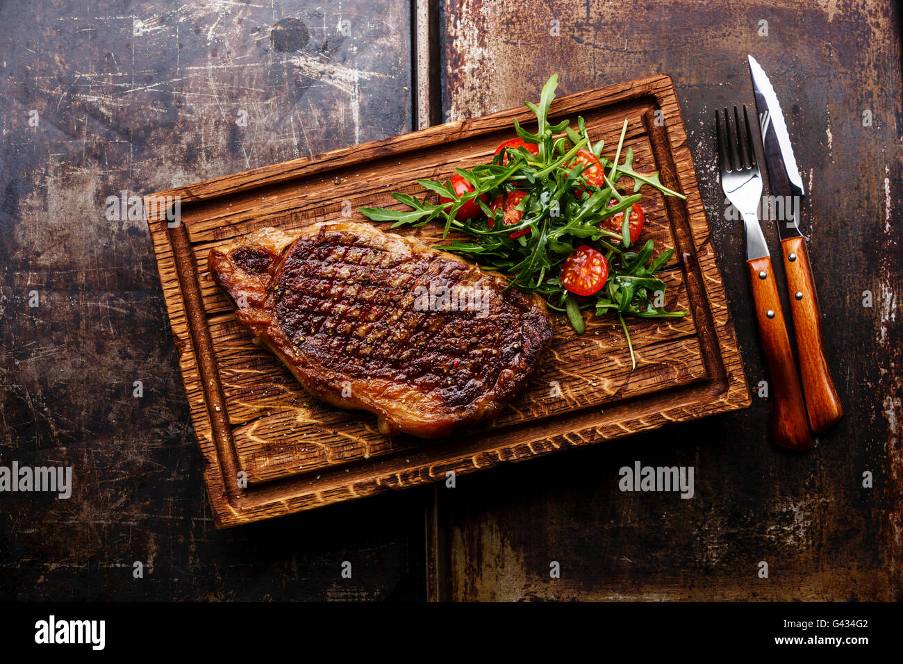 Alla brace bistecca di controfiletto con insalata e una forchetta e coltello sul tagliere su sfondo scuro Foto Stock