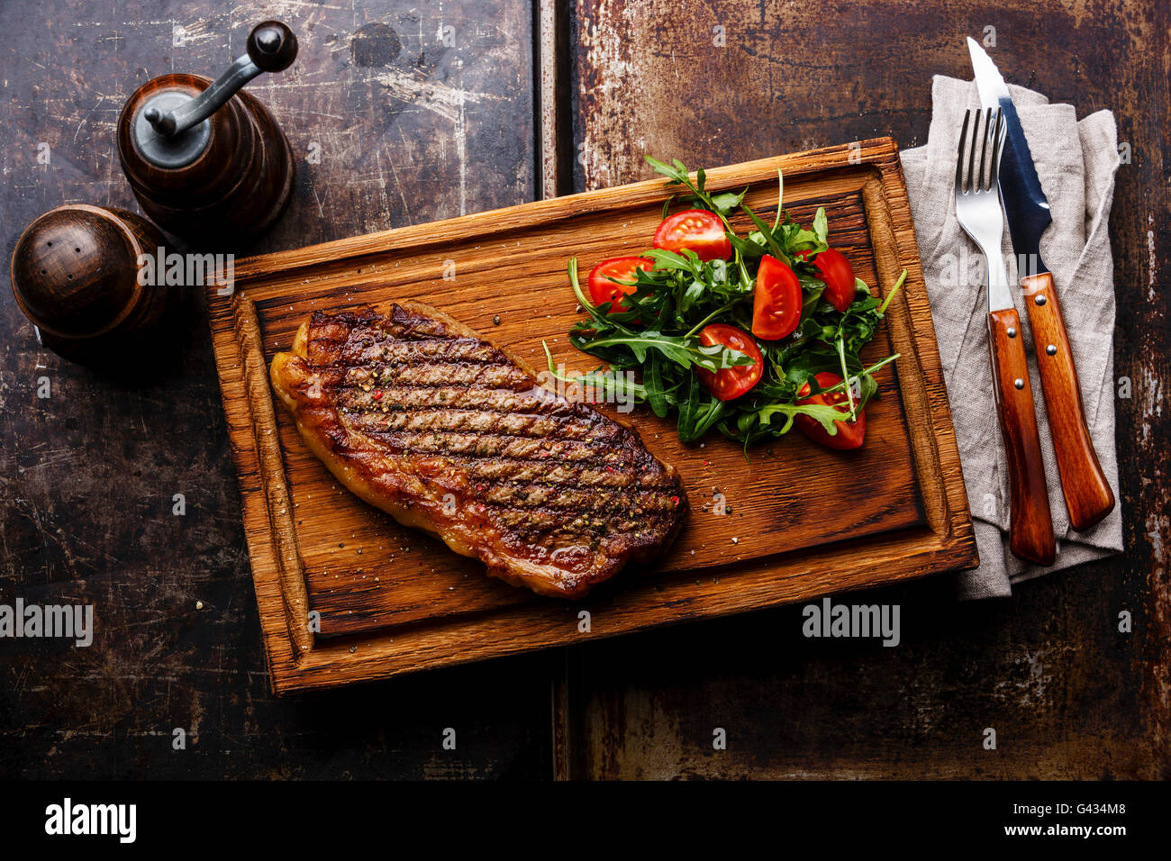 Alla brace bistecca di controfiletto con insalata e una forchetta e coltello sul tagliere su sfondo scuro Foto Stock