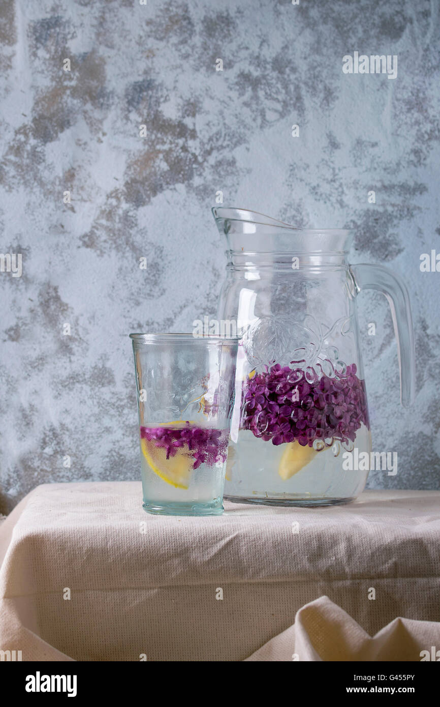 Il vetro e la brocca di limonata lilla Acqua con limone e fiori lilla, servita sulla biancheria da letto bianca tovaglia con blue textured wal Foto Stock