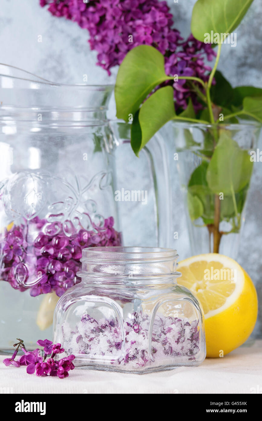 Il vasetto di vetro di lilla fiori in zucchero, il vetro e la brocca di lillà Acqua con limone e il ramo di lilla fresca sulla biancheria da letto bianca tabella Foto Stock