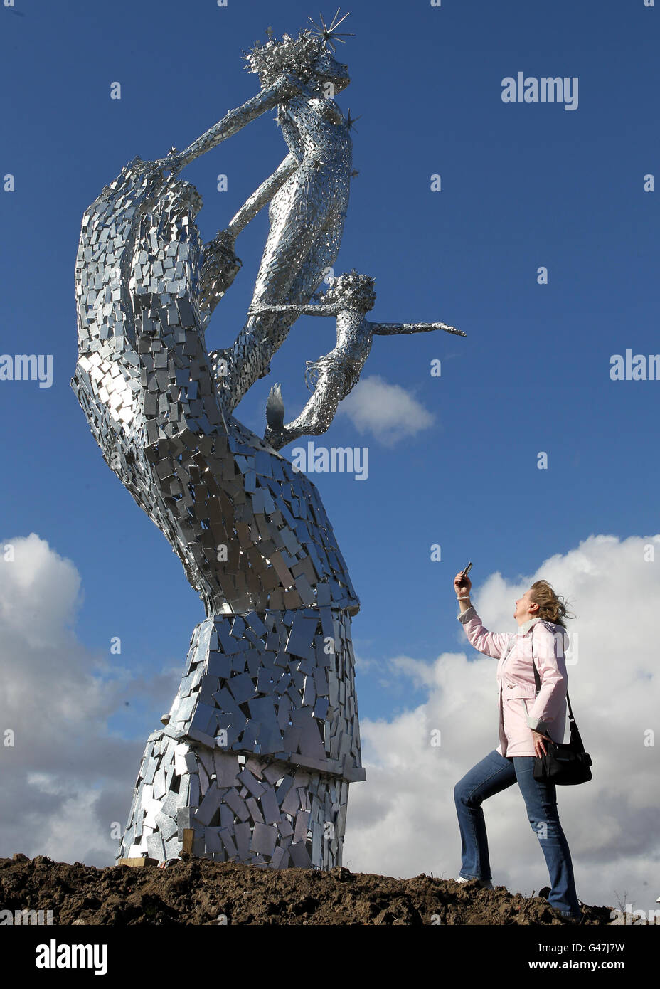 Una donna scatta una foto sul suo telefono di una scultura in acciaio alta 8 m di una mano gigante che sostiene una donna e un bambino di nome 'Lifeline' dall'artista Andy Scott, ispirato dal lavoro dei servizi di emergenza, installato oggi alla rotonda di Shillinghill in Alloa. Foto Stock