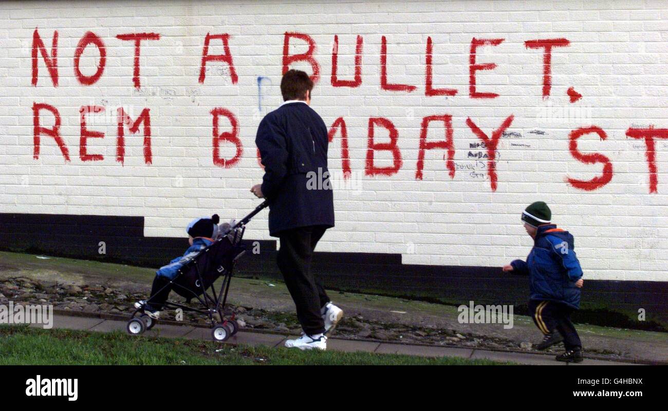 Una madre con i suoi figli passa per graffiti a Belfast ovest facendo riferimento alla linea di smantellamento e a un ricordo di quanto accaduto a Bombay Street a Belfast nel 1969, quando i Loyalists attaccarono e bruciarono le case cattoliche. Foto Stock