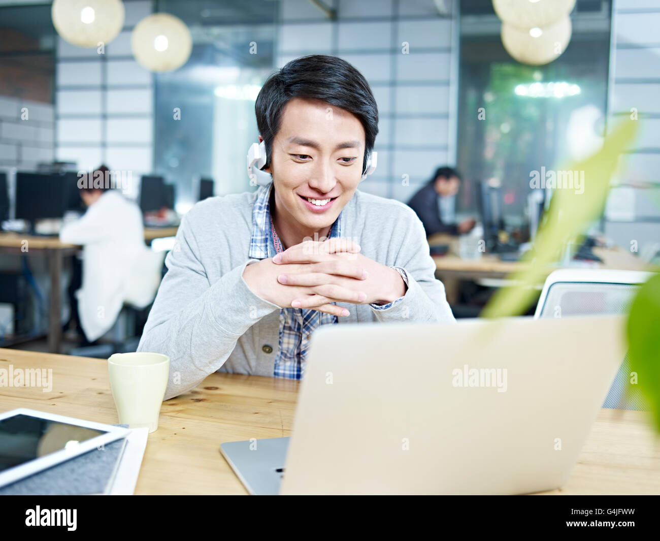 Giovani designer asiatici guardando il computer portatile durante l'ascolto di musica con cuffie in ufficio. Foto Stock