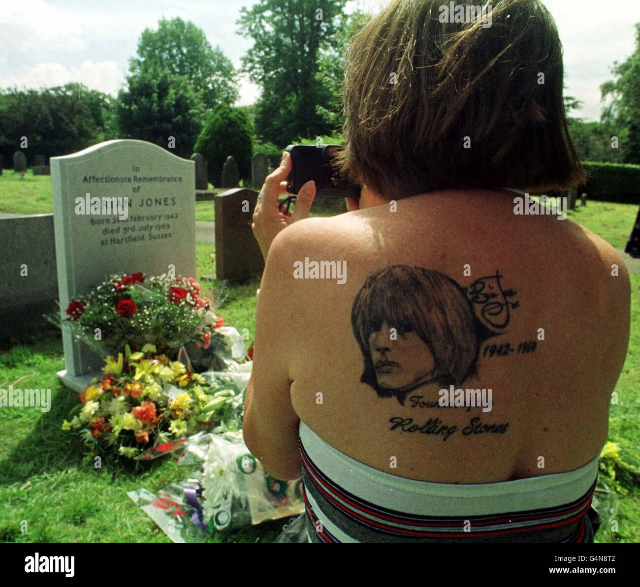 Margie Calvano di 50 anni, dal New Jersey, USA, con un tatuaggio Brian Jones sulla spalla, scatta una fotografia alla tomba di Cheltenham, della stella Rolling Stones che morì 30 anni fa. Foto Stock
