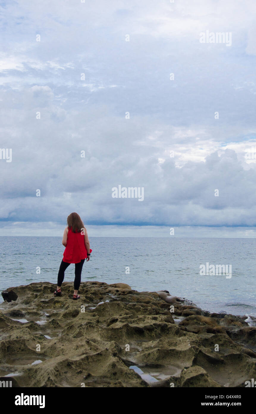 Ragazza in rosso che affaccia sulla spiaggia rocciosa di oceano su nuvoloso giorno vicino al tramonto Foto Stock