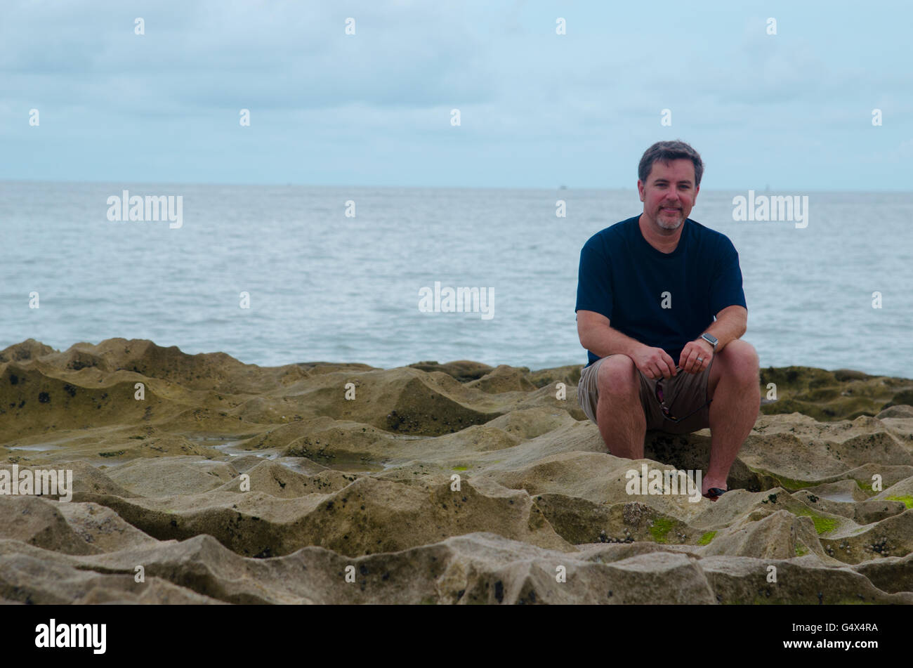 Orizzontale dell uomo sorridente seduto sulla spiaggia rocciosa contro il cielo al tramonto indossa watch Foto Stock