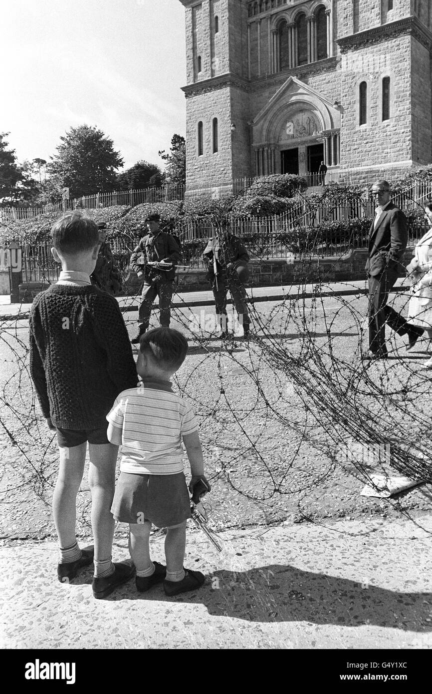 Uno dei cittadini più piccoli di Belfast porta la sua pistola giocattolo mentre guarda i soldati armati attraverso una barricata a filo spinato sulla Crumlin Road. Sullo sfondo si trova la Cappella Ardoyne. Foto Stock
