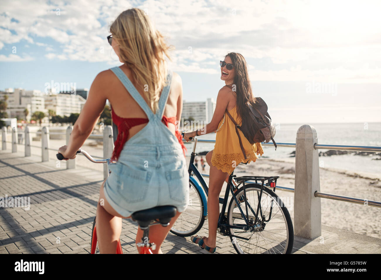 Vista posteriore di due giovani donne in bicicletta lungo la passeggiata sul lungomare. Migliori amici a cavallo sulle loro moto in riva al mare Foto Stock