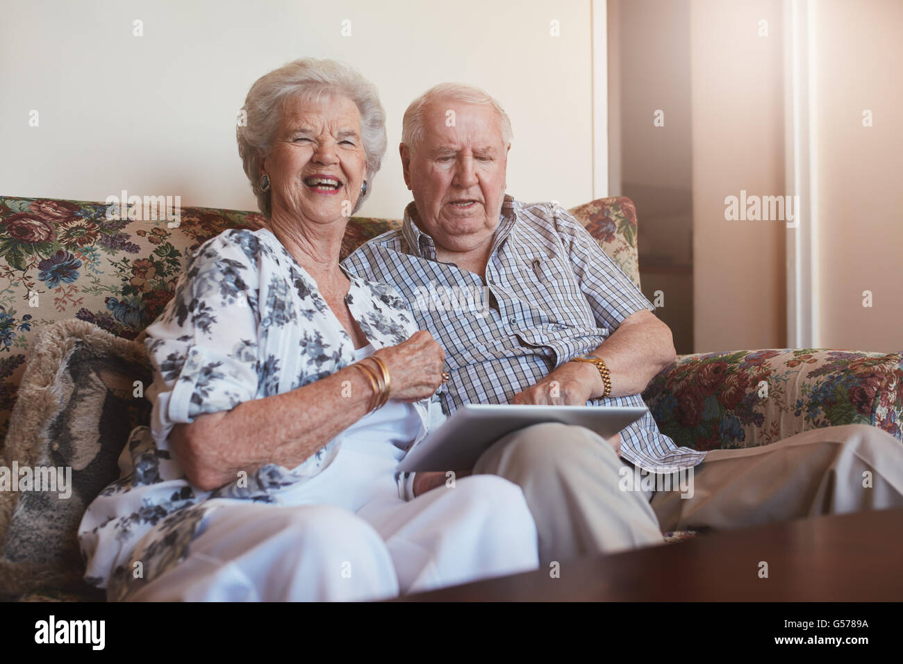 Ritratto di sorridere senior donna seduta con il marito usando digitale compressa. Coppia di anziani seduti sul divano di casa con un t Foto Stock
