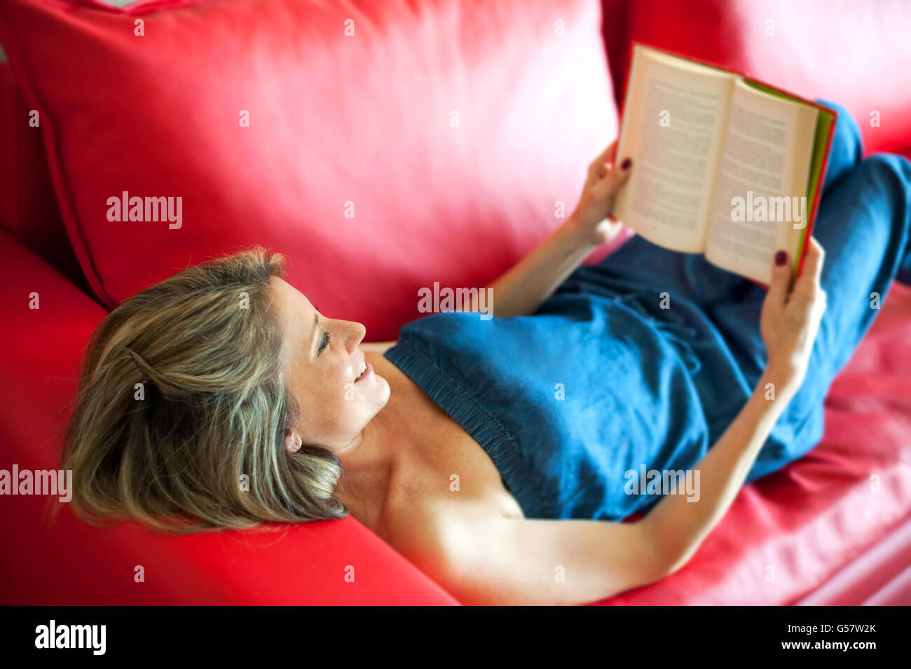 Attraente donna matura sorridente in godimento come lei legge un libro mentre un momento di relax a casa su un rosso di divano in pelle Foto Stock