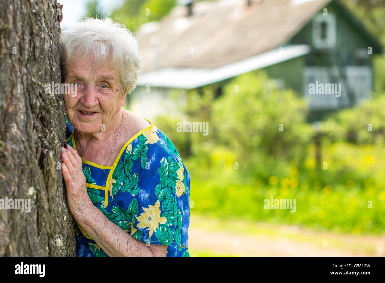 Una donna anziana sorge nei pressi di un albero con casa rurale in background. Foto Stock