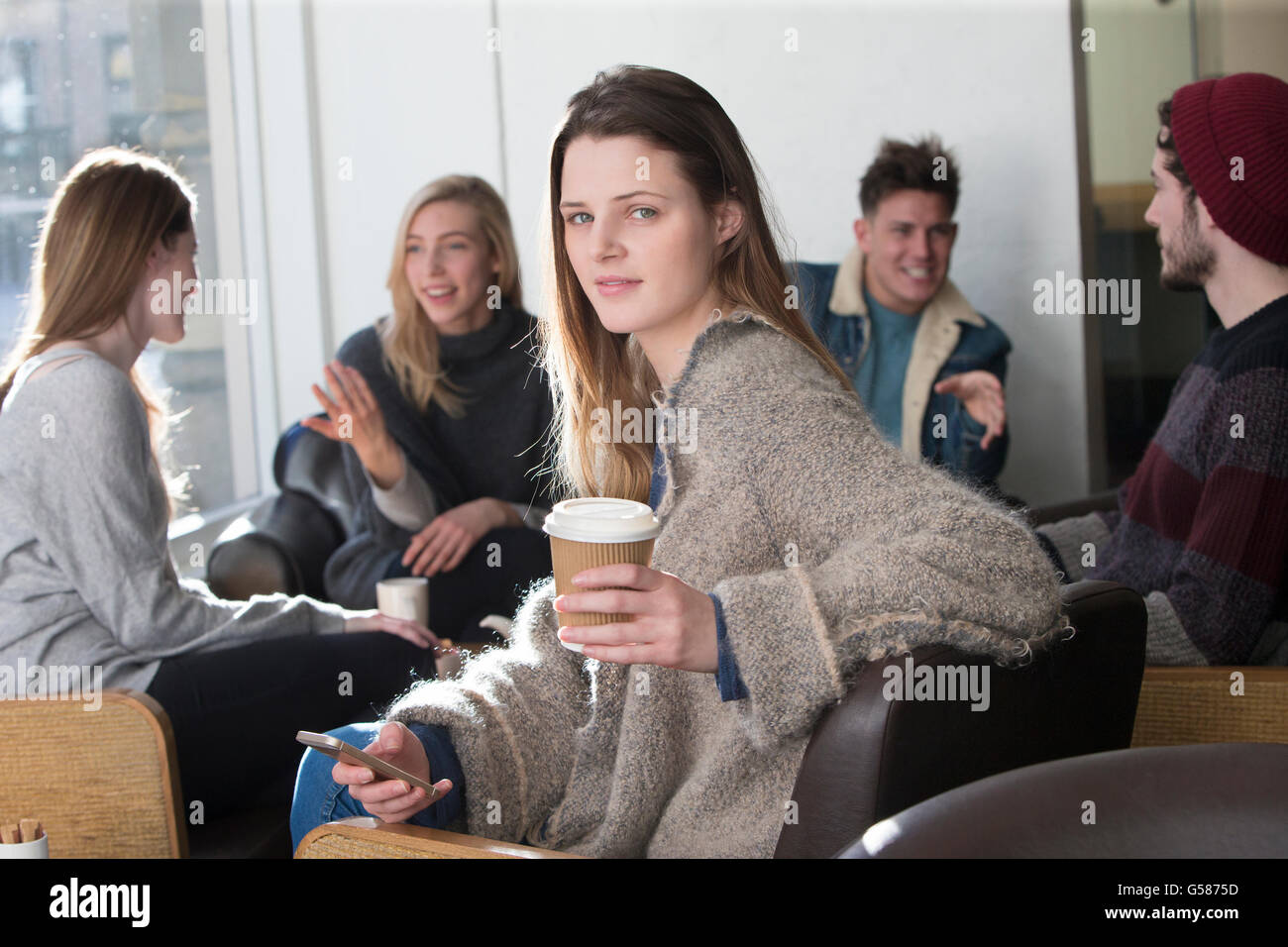 Giovane donna sorridente per la telecamera in un bar con i suoi amici in background. Ella è in possesso di uno smartphone e un bicchiere di COF Foto Stock