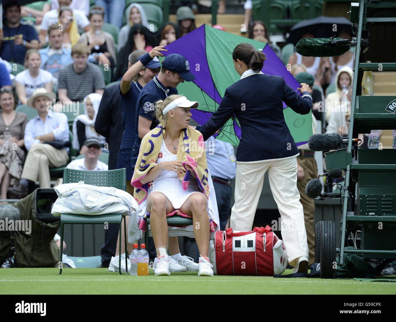Il danese Caroline Wozniacki attende come un ragazzo di palla e l'umpire tenta di aprire un ombrello durante il terzo giorno dei Campionati di Wimbledon 2012 presso l'All England Lawn Tennis Club di Wimbledon. Foto Stock