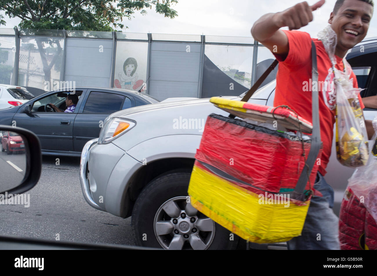Benvenuto a Rio - strada sorridente venditore lavora durante le ore di punta di traffico a Linha Vermelha expressway ( Linea Rossa ), il principale collegamento tra il Rio de Janeiro / Antonio Carlos Jobim International Airport e la città, Brasile - opache in plastica dura e la divisione tra la strada e la Favela da mare, zona dominata dal traffico di stupefacenti. Foto Stock