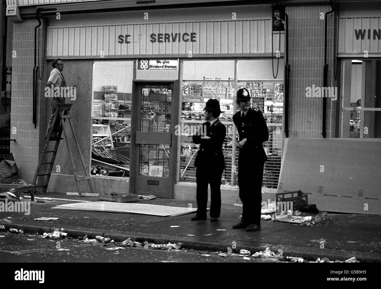 NOTTING HILL 1976: Guardia di stand della polizia come un addetto al lavoro sale le finestre del supermercato Co-op e fuori licenza a Westbourne Park Road seguendo le scene di violenza della scorsa notte a Notting Hill, Londra ovest, quando un carnevale indiano occidentale è esploso in una rivolta. Foto Stock