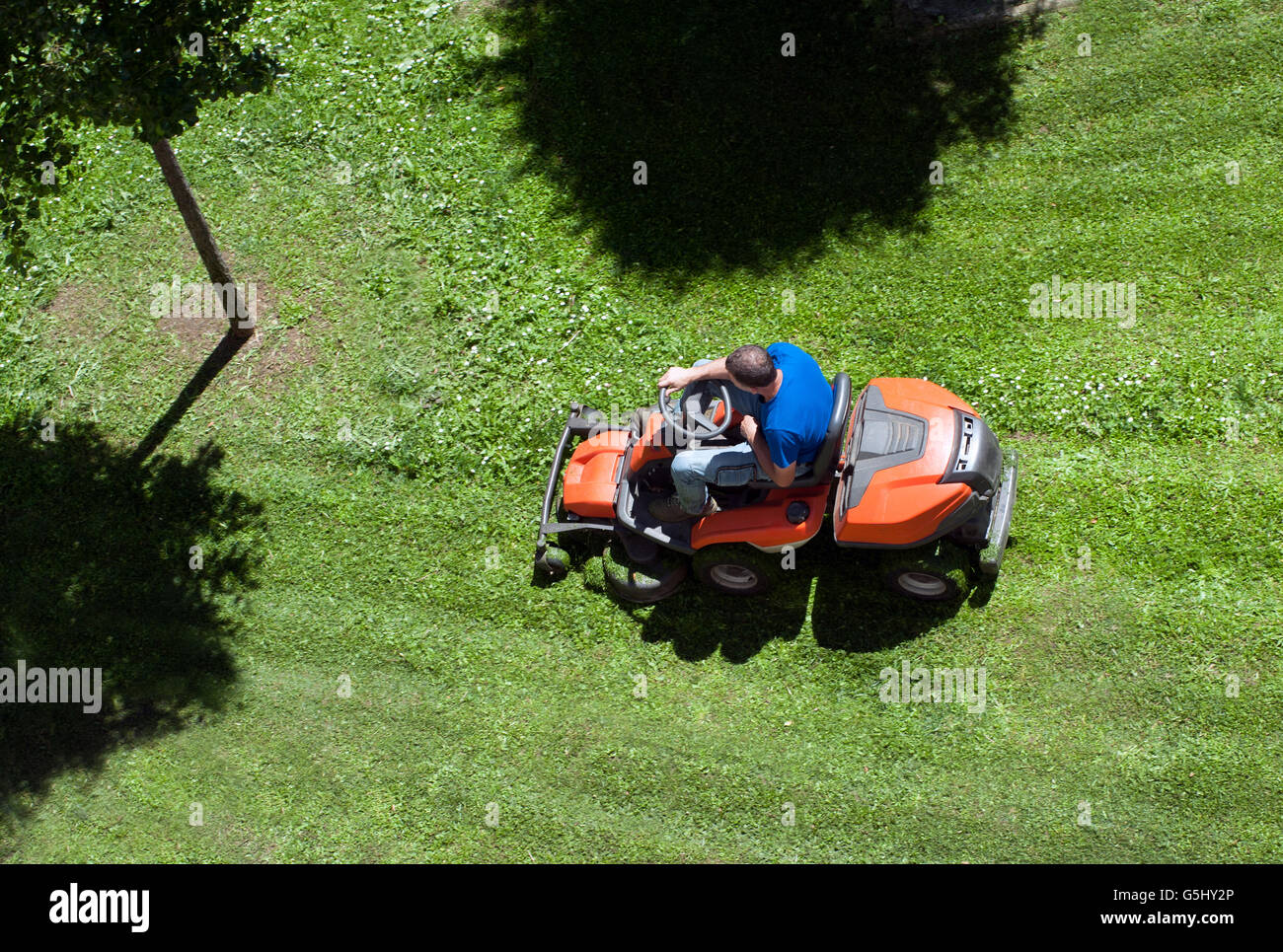 Vista aerea di un uomo di falciare un prato su un arancio ride su tosaerba con le ombre degli alberi in un concetto di manutenzione di cantiere Foto Stock