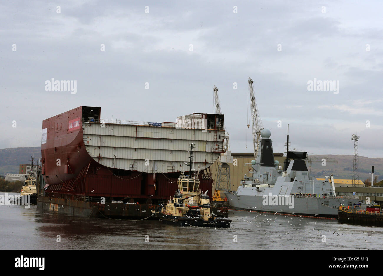 Un'enorme sezione di una nuova portaerei della Royal Navy, la sezione di 11,300 tonnellate di scafo a poppa per la HMS Queen Elizabeth, è portata da una chiatta di mare a circa 600 miglia da Govan, Glasgow, a Rosyth sul Firth of Forth, passando una nave da guerra in un altro molo. Foto Stock