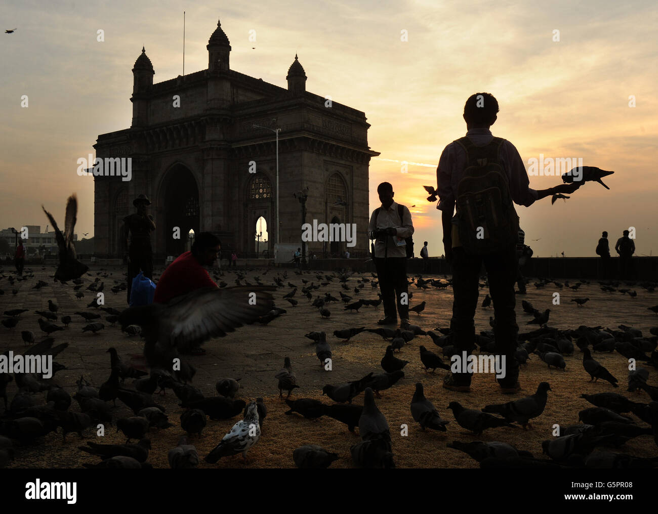 Un uomo alimenta piccioni come il sole sorge dietro la porta d'India nella zona Apollo Bunder di Mumbai, India PRESS ASSOCIATION foto. Data immagine: Giovedì 29 novembre 2012. Costruito per commemorare la visita di re Giorgio V e della regina Maria a Mumbai nel dicembre 1911 (tuttavia, hanno visto solo un modello di cartone della struttura, dal momento che la costruzione non è iniziata fino al 1915) Le ultime truppe britanniche a lasciare l'India in seguito all'indipendenza dell'India passarono attraverso il gateway in una cerimonia il 28 febbraio 1948, segnalando la fine del governo britannico in India (Raj britannico). Il credito fotografico dovrebbe essere: Anthony Foto Stock