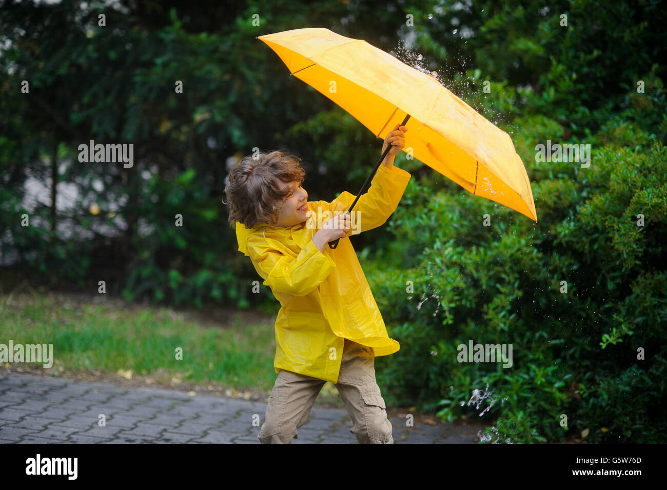 Ragazzino in un impermeabile giallo è coperto con un ombrello da pioggia e vento. Vento sfrangiature il ragazzo del capello. Egli difficilmente detiene una grande y Foto Stock