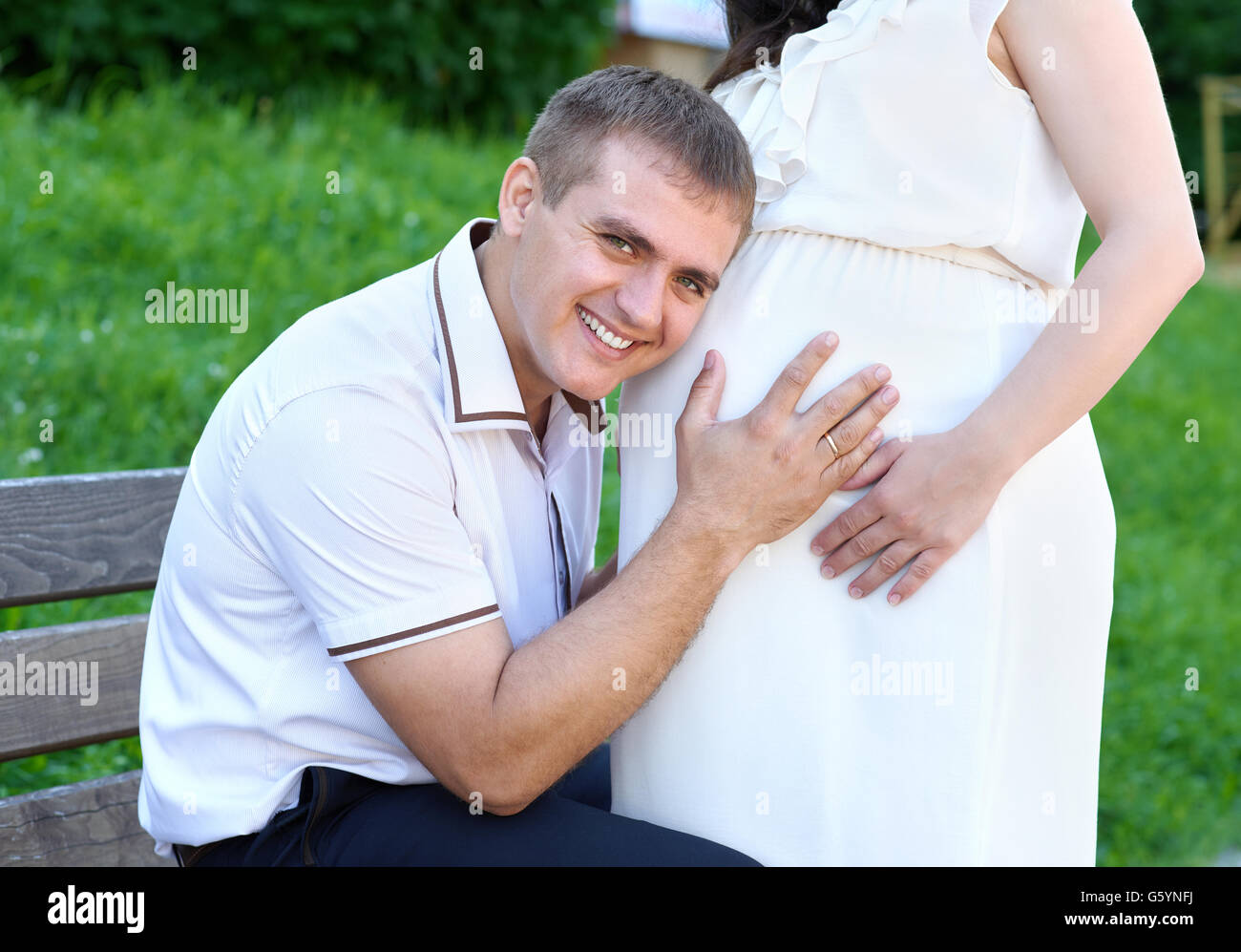 La donna incinta e il marito, uomo pancia ascoltare e parlare con il bambino e la famiglia felice, giovane nel parco cittadino, durante la stagione estiva, erba verde un Foto Stock