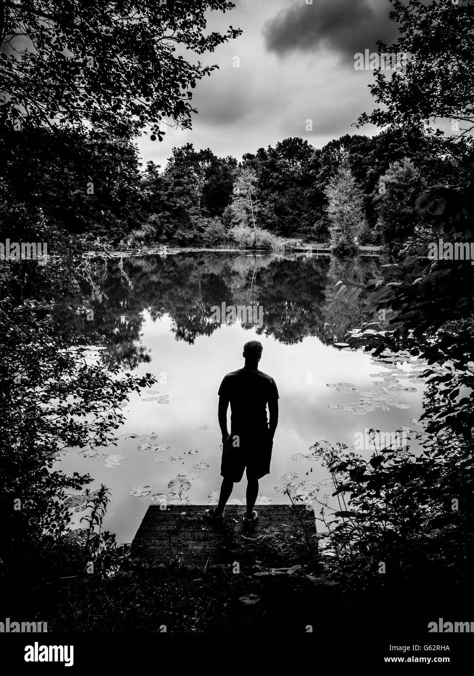 Silhouette di uomo si trovava su un molo di legno che si affacciava sul lago con gli alberi che circondano i bordi Foto Stock