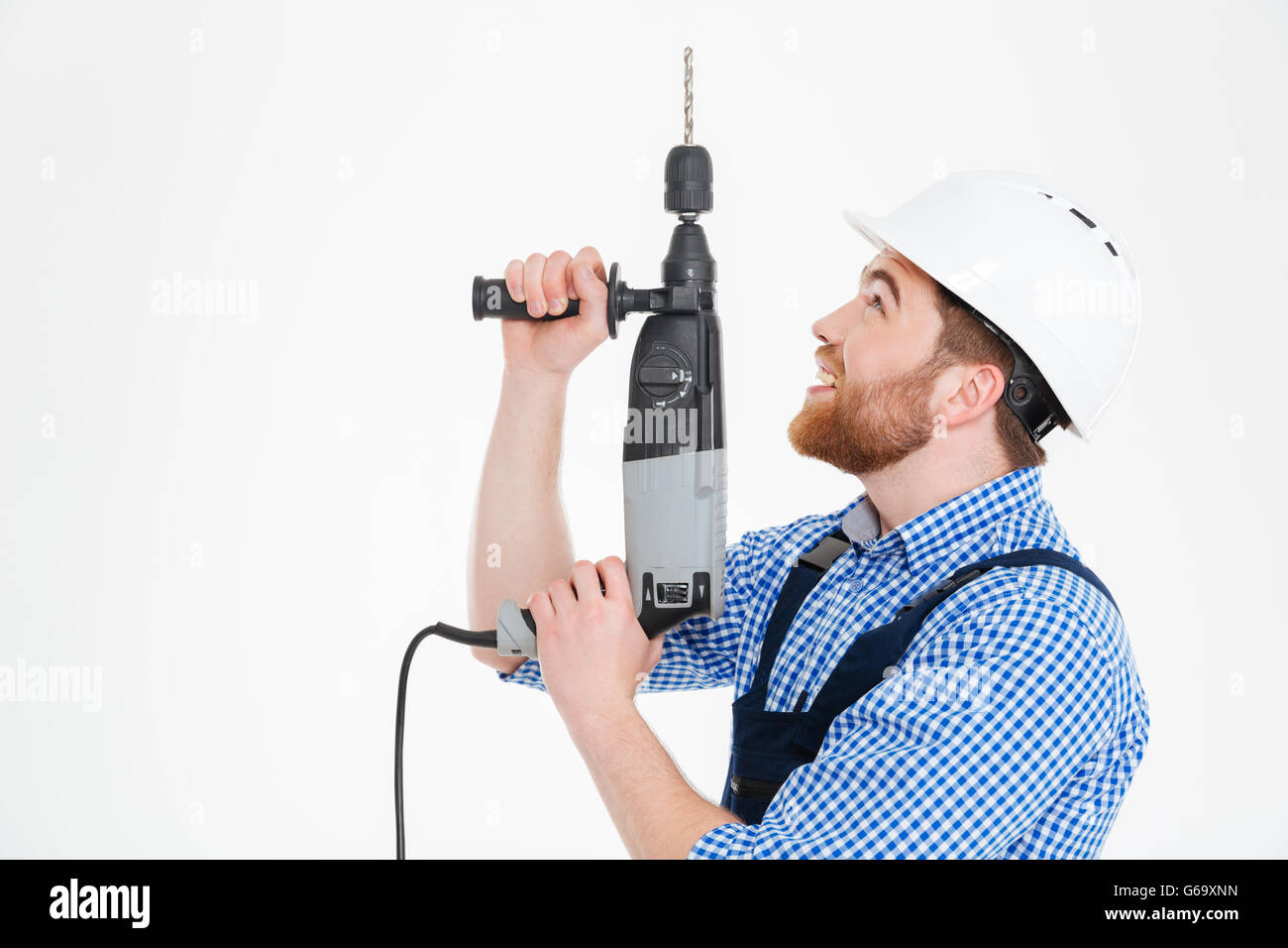 Sorridente giovane barbuto builder nel casco mediante trapano e guardando in alto Foto Stock