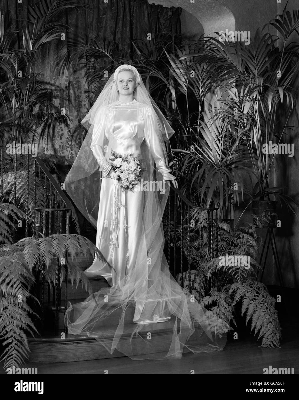 1930s ritratto a piena lunghezza sposa bouquet di contenimento che indossa velo Abito in raso tra le palme e le felci in ambienti interni Foto Stock