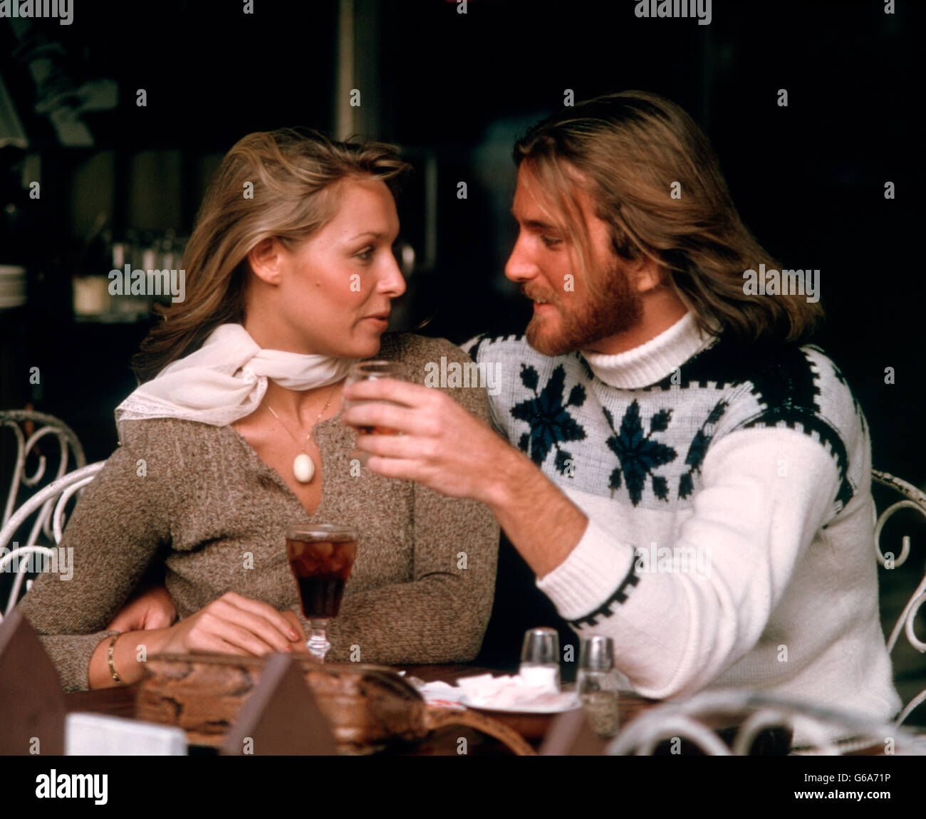 Anni Settanta coppia romantica uomo donna con drink seduti in Outdoor Cafe entrambi indossando maglioni uomo ha i capelli lunghi e la barba Foto Stock