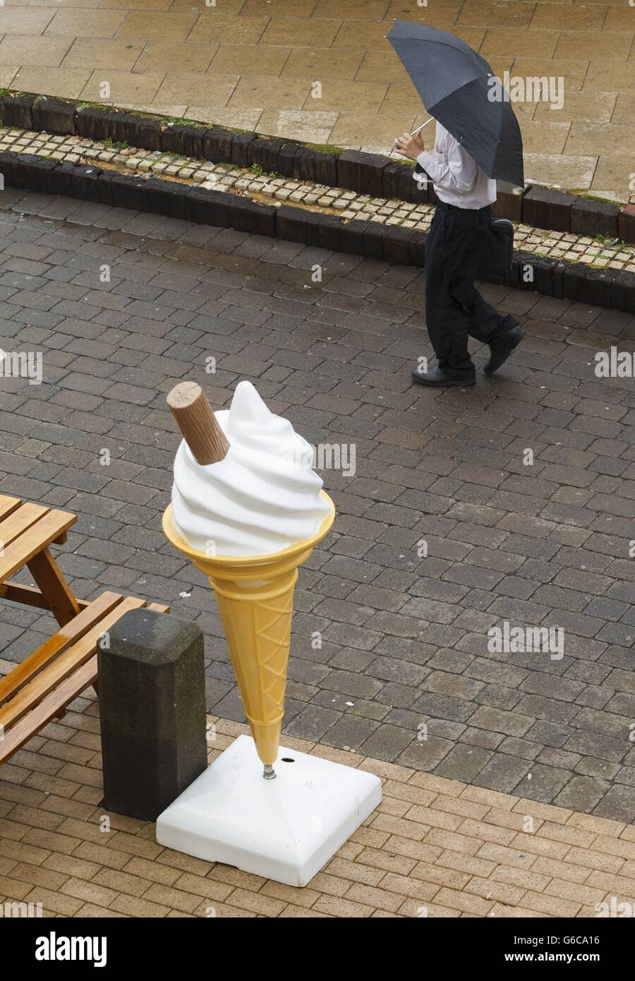 Un uomo sotto un ombrello cammina davanti a un gigantesco gelato di plastica sulla spiaggia di Brighton, East Sussex, all'inizio del weekend di agosto Bank Holiday. Foto Stock
