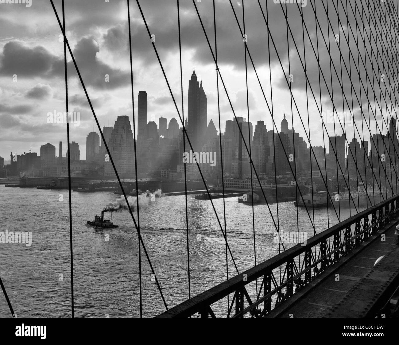 Negli anni quaranta anni cinquanta dello skyline di downtown Manhattan visto attraverso i cavi del ponte di Brooklyn Bridge RIMORCHIATORE IN EAST RIVER NEW YORK NY USA Foto Stock