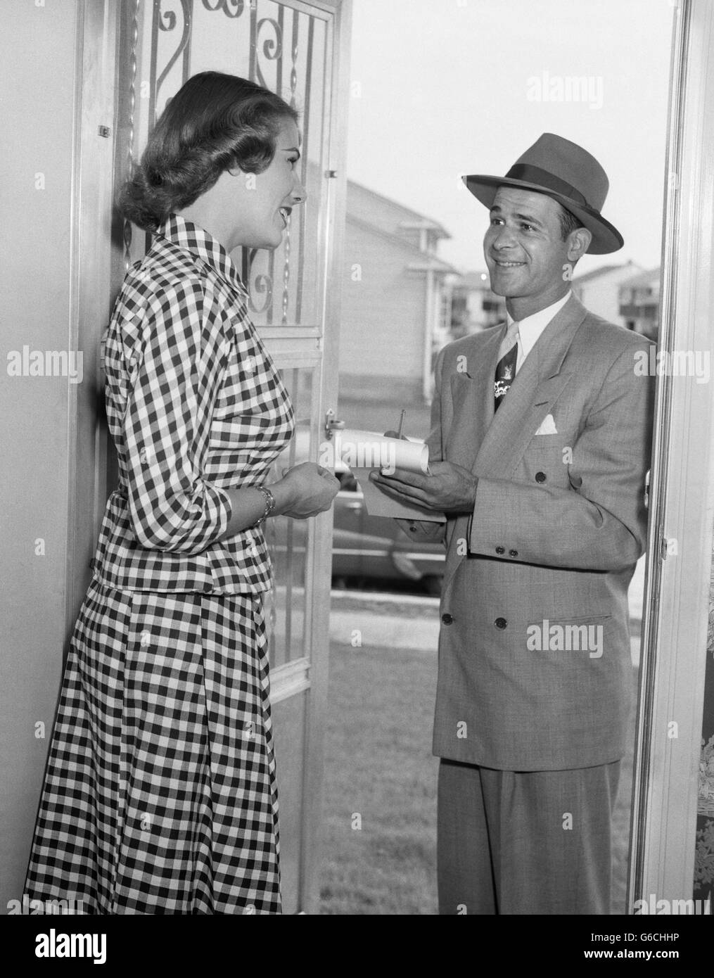 Anni Cinquanta massaia IN ABITO GINGHAM presso la porta di casa parlando AL VENDITORE IN TUTA & HAT HOLDING matita e PAD Foto Stock