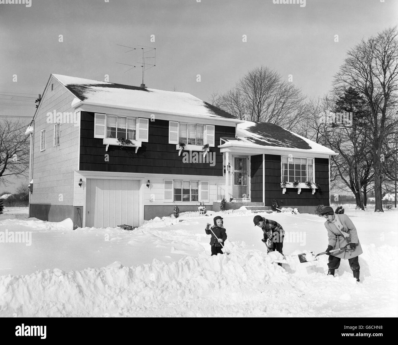 Degli anni Cinquanta e madre di 2 figli di fronte casa spalare la neve profonda da marciapiede e carraio Foto Stock