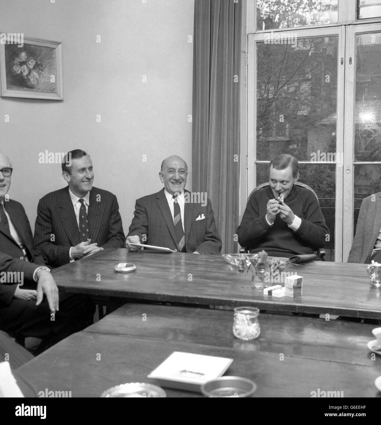 Vestito informalmente con un maglione blu, il sig. ANTHONY WEDGWOOD BENN, il Postmaster-General, accende il suo tubo all'inizio sorridente dei colloqui nel soggiorno della sua casa di Londra in Holland Park Avenue, Nottinghill, Su accordi televisivi fatti per il campionato del mondo lotta tra Cassius Clay e Henry Cooper a Londra il 21 maggio. Con lui nella foto sono l'ex capitano di cricket inglese TED DEXTER, un direttore esecutivo di Viewsport Ltd., e il sig. HARRY LEVENE, il promotore di lotta. Ai colloqui hanno partecipato anche rappresentanti della BBC, dell'ITV e della Pay Television. Foto Stock