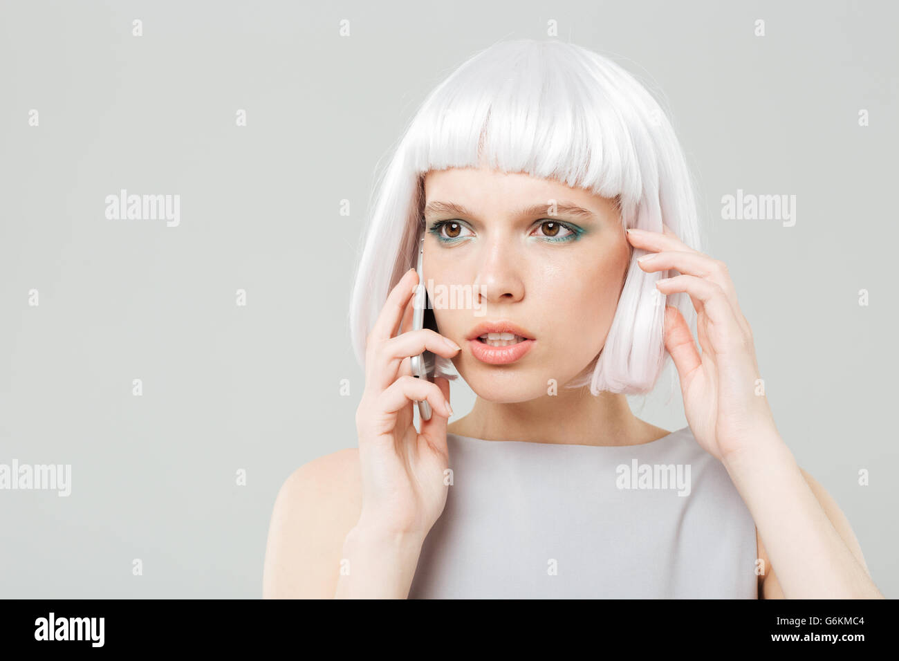Riflessivo preoccupato giovane donna in parrucca bionda parlando al telefono cellulare su sfondo bianco Foto Stock