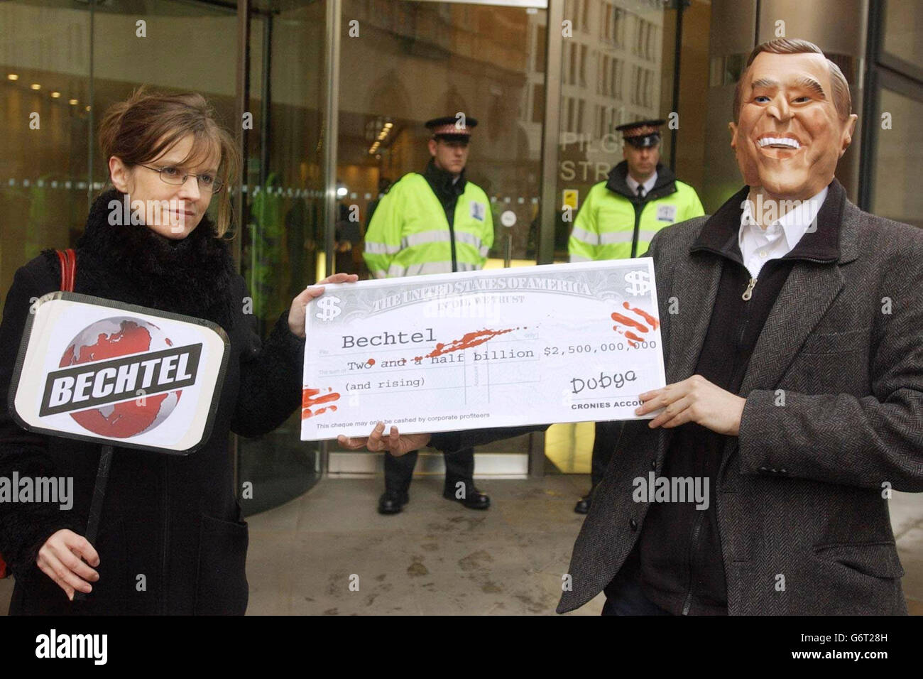 Un protetore vestito come il presidente americano George W Bush tiene un controllo falso impaurito con 'il sangue' fuori degli uffici londinesi di Bechtel. I manifestanti stavano dimostrando di quello che chiamano l'invasione aziendale dell'Iraq. Foto Stock