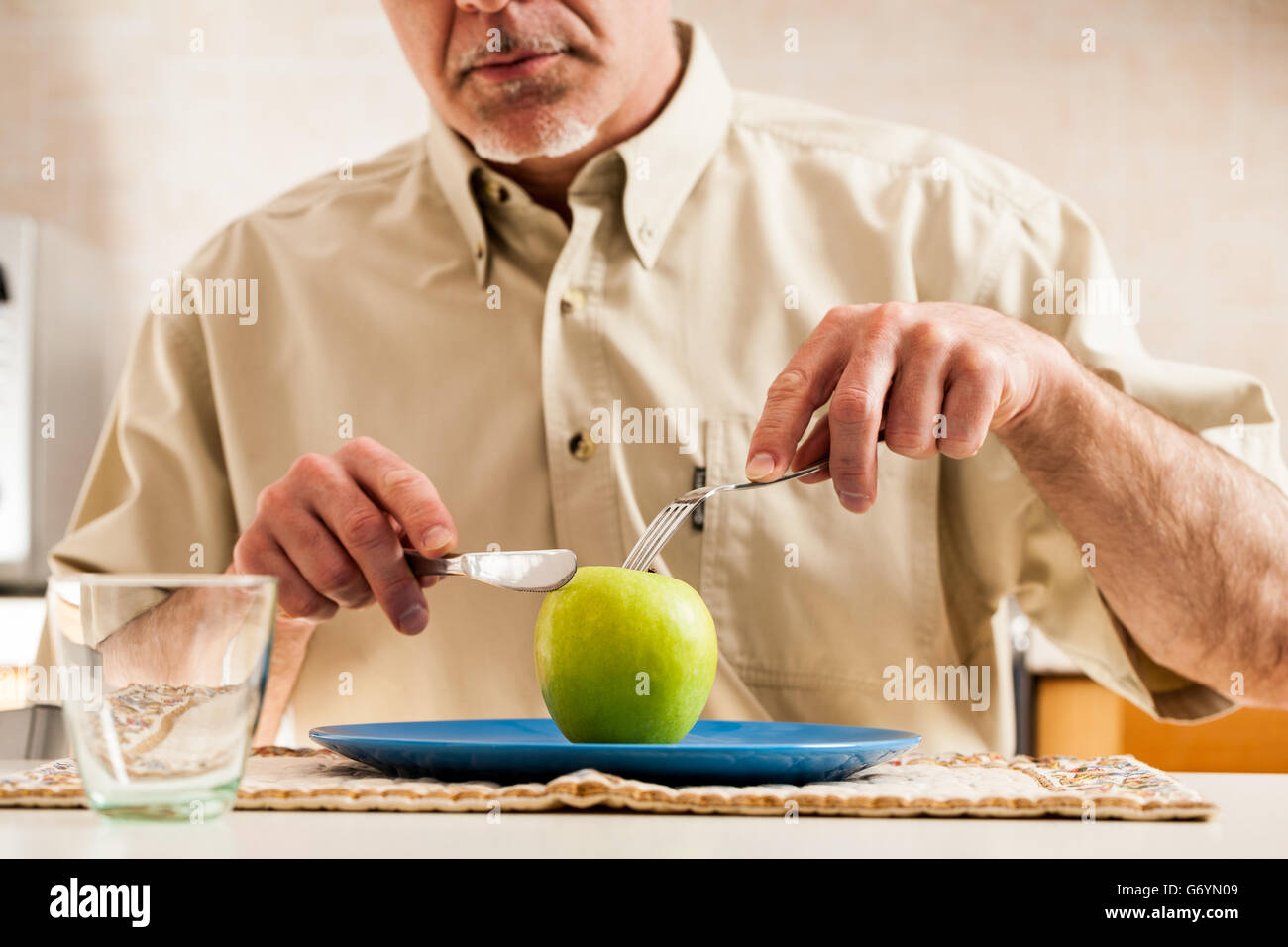 Oscurata l Uomo in camicia marrone con coltello e forchetta per affettare e mangiare tutta una mela verde sulla parte superiore della piastra di blu Foto Stock