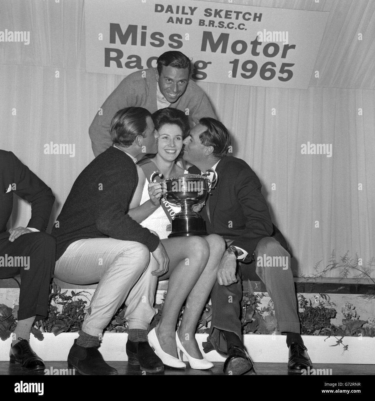 Jackie Peterson, di Saltdean, Brighton, detiene il trofeo dopo essere stato eletto 'Miss Motor Racing 1965' a Brands Hatch, Kent. Si congratula con lei per i baci dei piloti di corse (l-r) Graham Hill, Jim Clark e Jack Brabham. Foto Stock