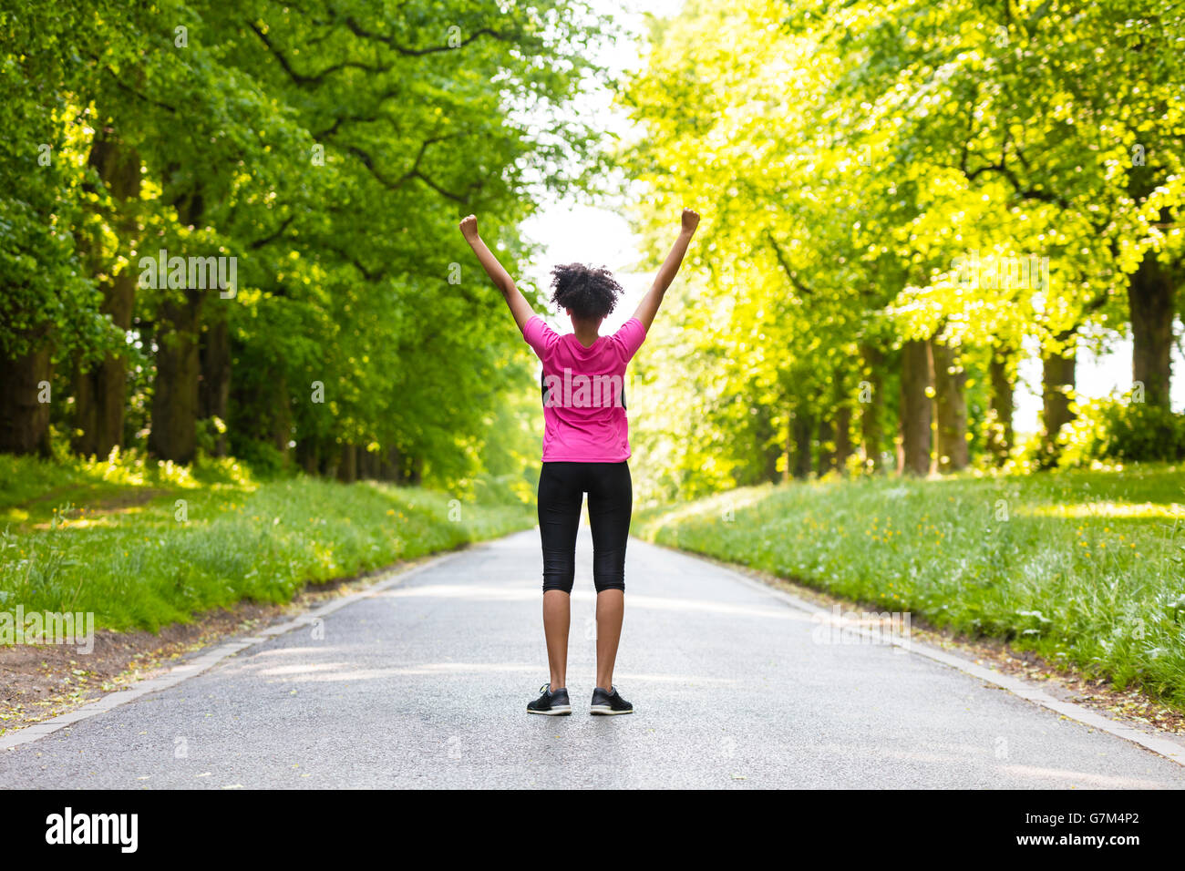Razza mista americano africano donna giovane ragazza adolescente fitness jogging in esecuzione per celebrare il successo sulla strada fiancheggiata con alberi verdi Foto Stock