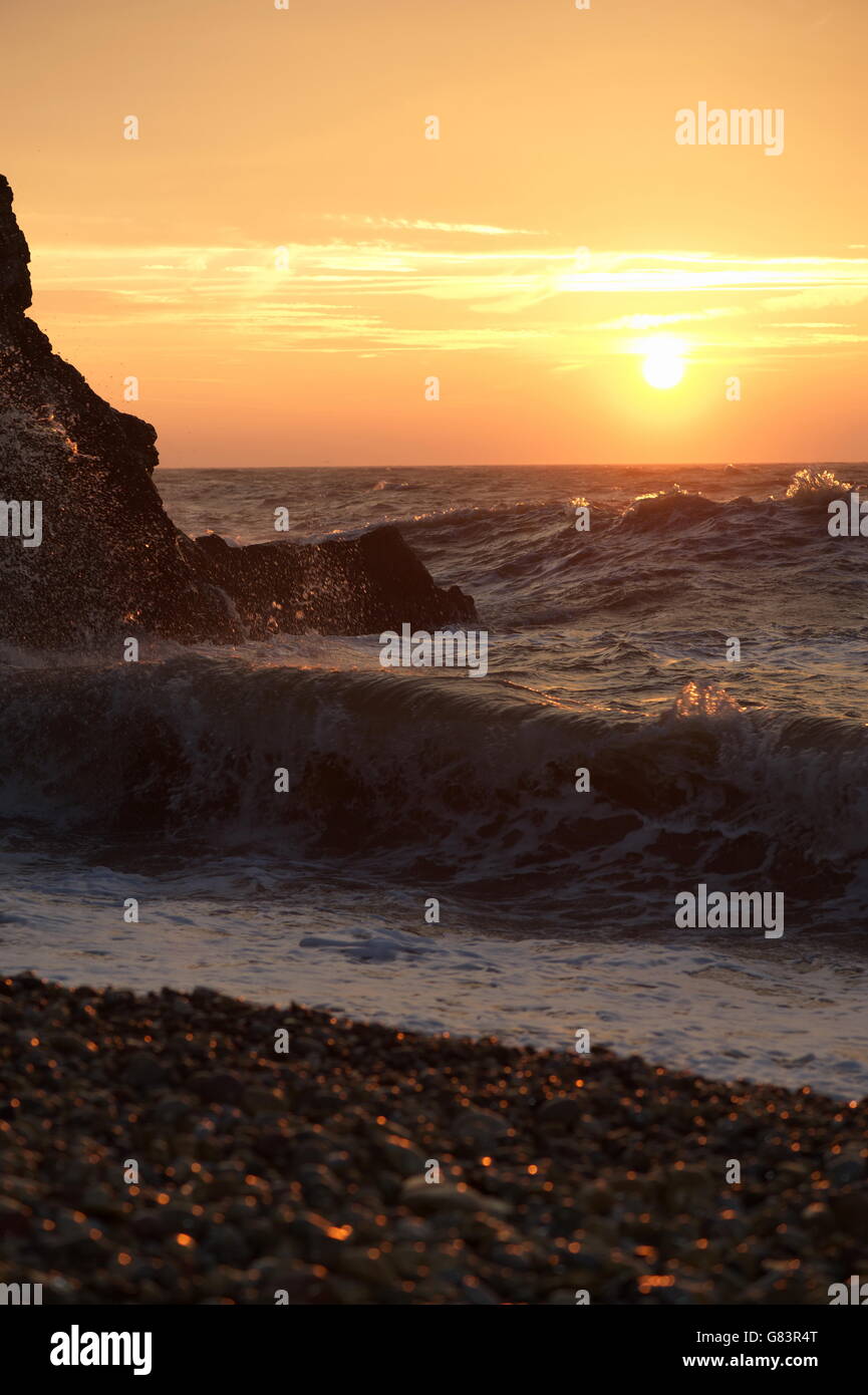 Le onde rompono su una riva di ghiaia a sunrise. Alba evidenzia lo spray e scogliere. Foto Stock